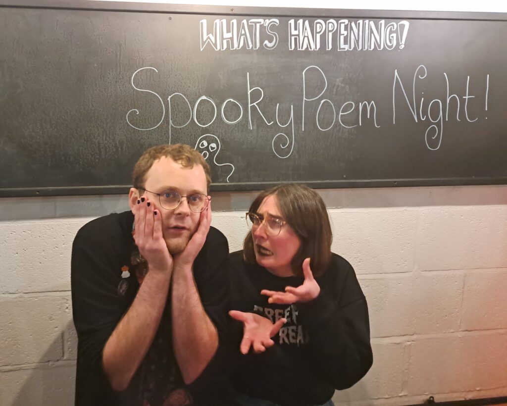 Lewis Brown (a white man with short brown hair, glasses, a black cardigan and nails painted black) and Megan Pattie (a white woman with brown hair, glasses, black lipstick and a shirt that says 'Creepin it Real') stand in mock panic in front a sign that says 'Spooky Poem Night'