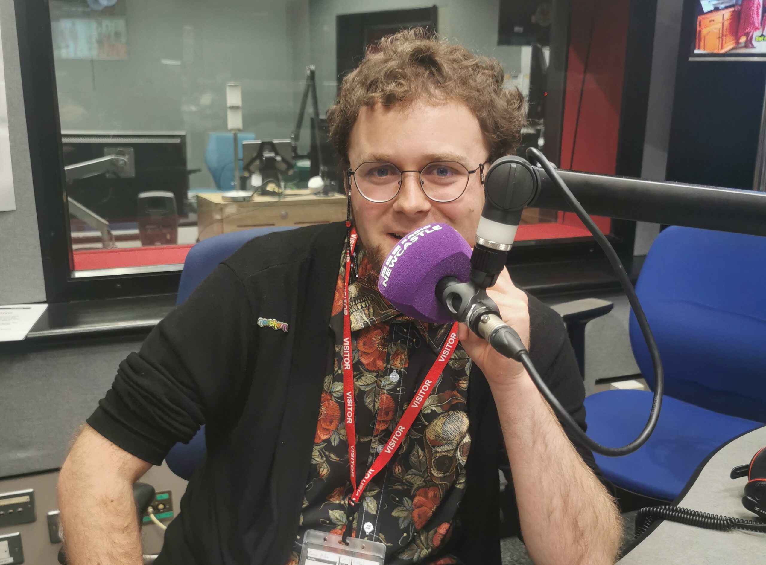 Lewis Brown (a young white man with curly hair and round glasses, wearing a black cardigan and a colorful shirt with roses and skulls) is speaking into a microphone with the words BBC Radio Newcastle written on it