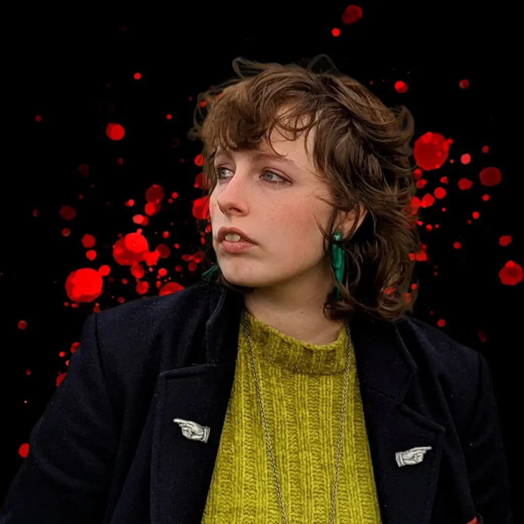 Kym Deyn is pictured (a white feminine person with long curly brown hair, a light blue earring, a black jacket and a yellow sweater). The background is black with red splotches, evocative of stylised bloodstains.