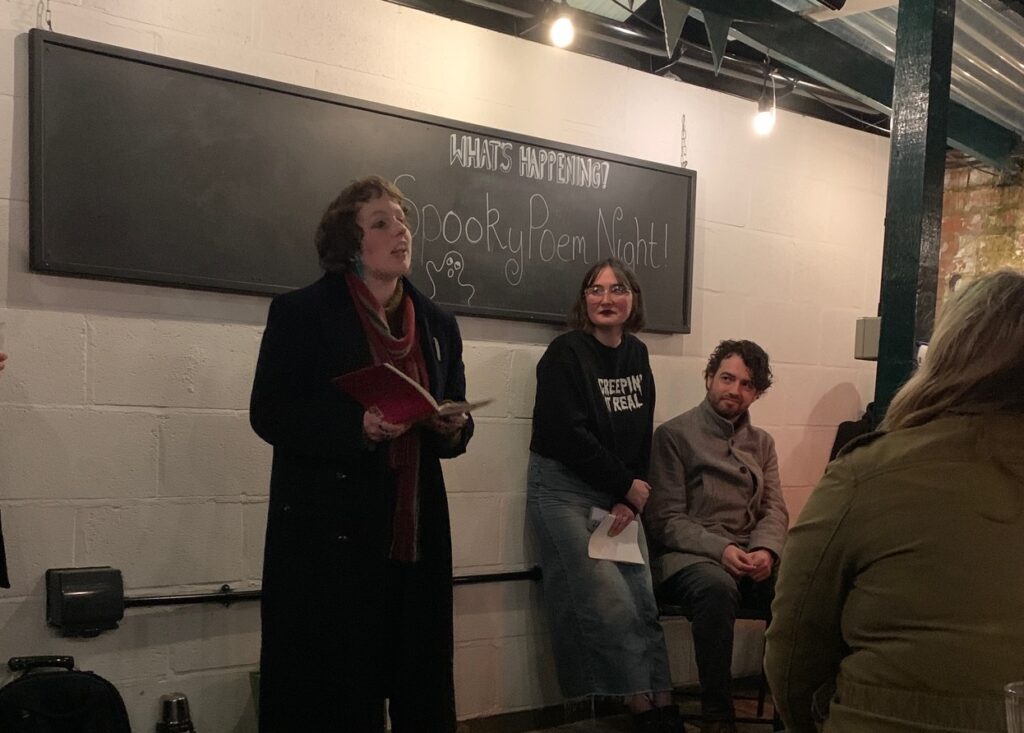 Kym Deyn (a white, feminine person wearing a black coat and a colorful scarf) stands in front of a sign reading 'Spooky Poem Night', and is reading poems from a red book