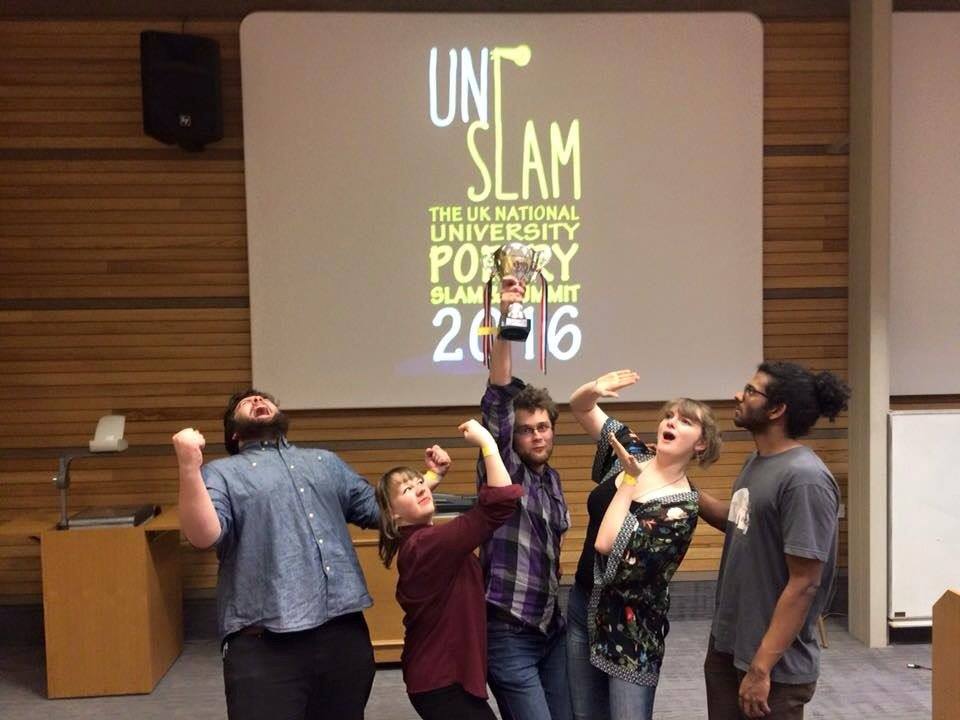 Five poets stand celebrating and holding up a trophy in front of a screen that says 'UniSlam: The UK National University Poetry Slam Summit 2016'.
