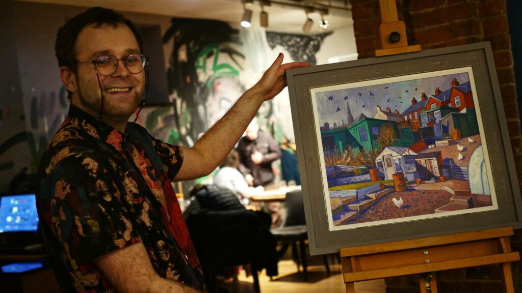 Poet Lewis Brown stands in a gallery posing with a colorful painting of some allotments by Chris Cyprus - the painting is called Racing Green