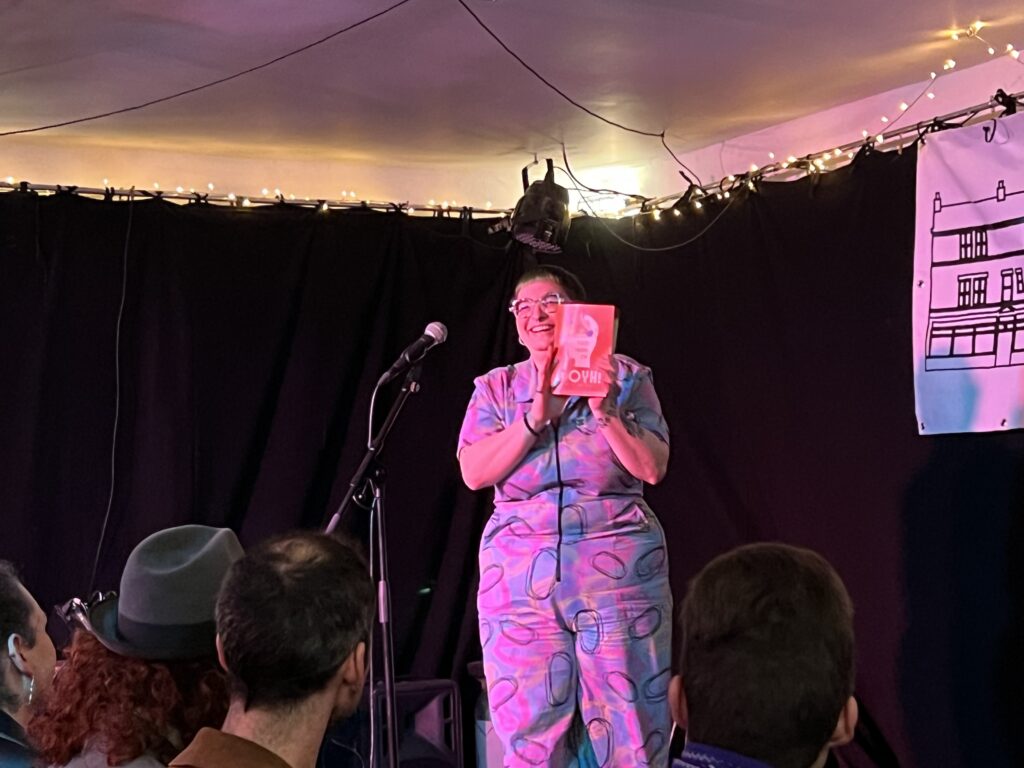 Poet Amy Langdown - with short hair, chunky glasses and a colorful boilersuit - stands on a stage holding a printed zine that says 'Out of Your Head'