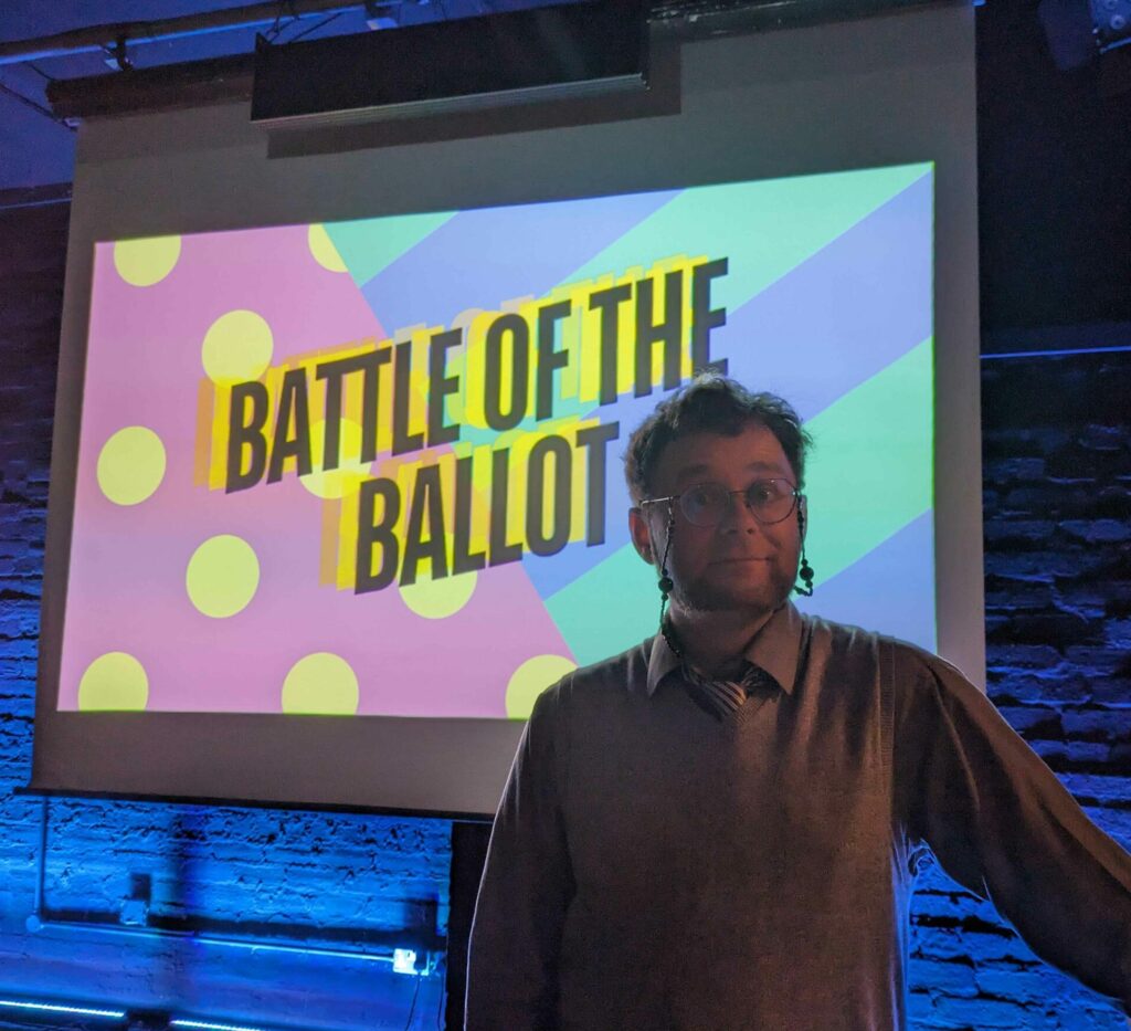 Poet Lewis Brown is dressed in a grey shirt and vest, and stands smiling in front of a projector screen that says Battle of the Ballot